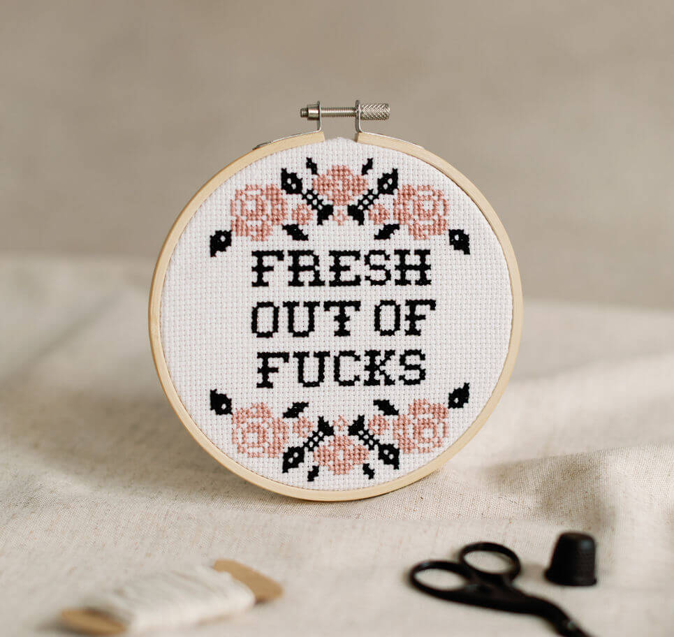 Embroidery hoop with adult cross-stitched text 'FRESH OUT OF FUCKS' on a beige background