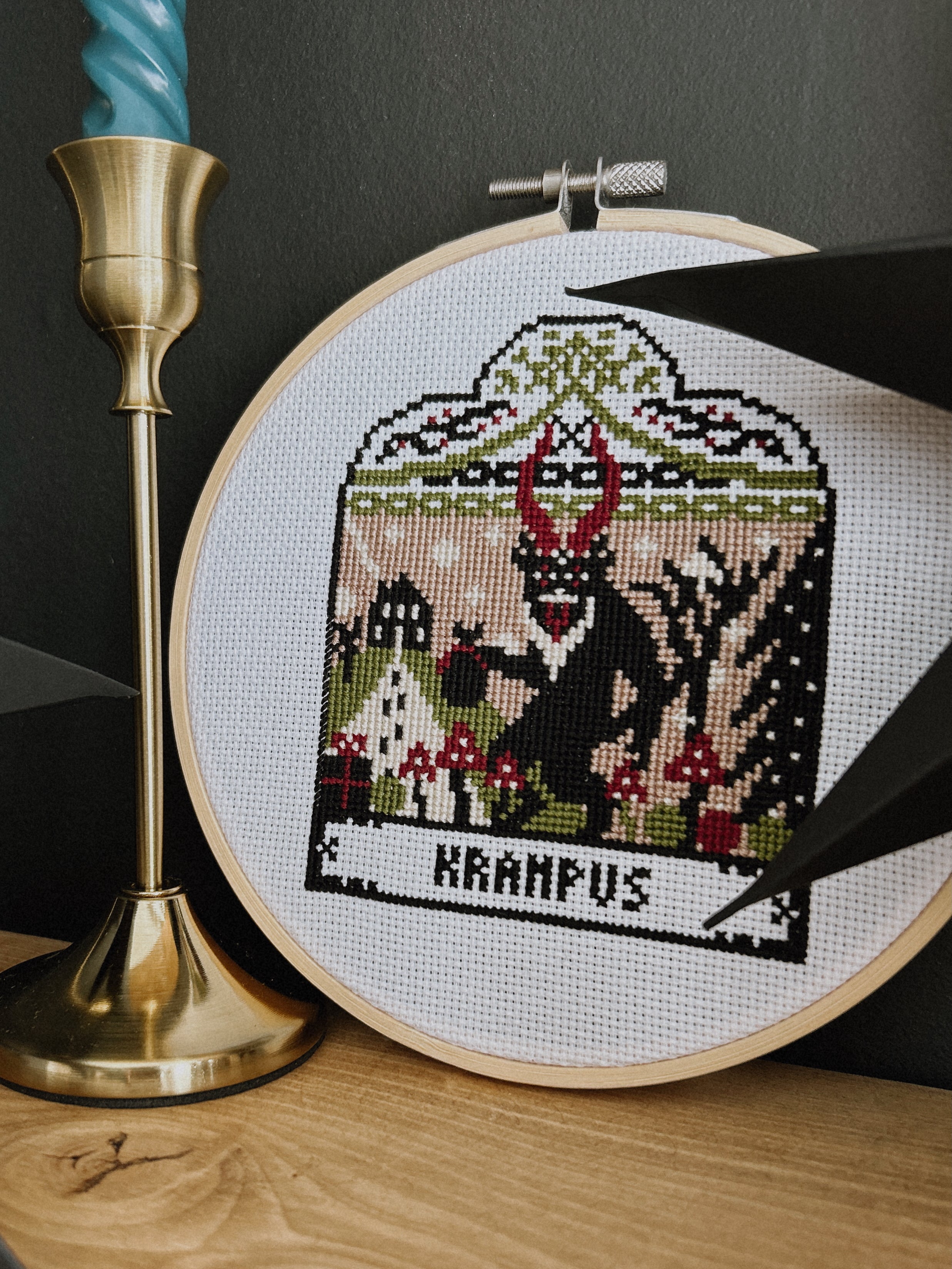 Cross-stitched design of Krampus in an embroidery hoop on a wooden surface with a candle in the background.